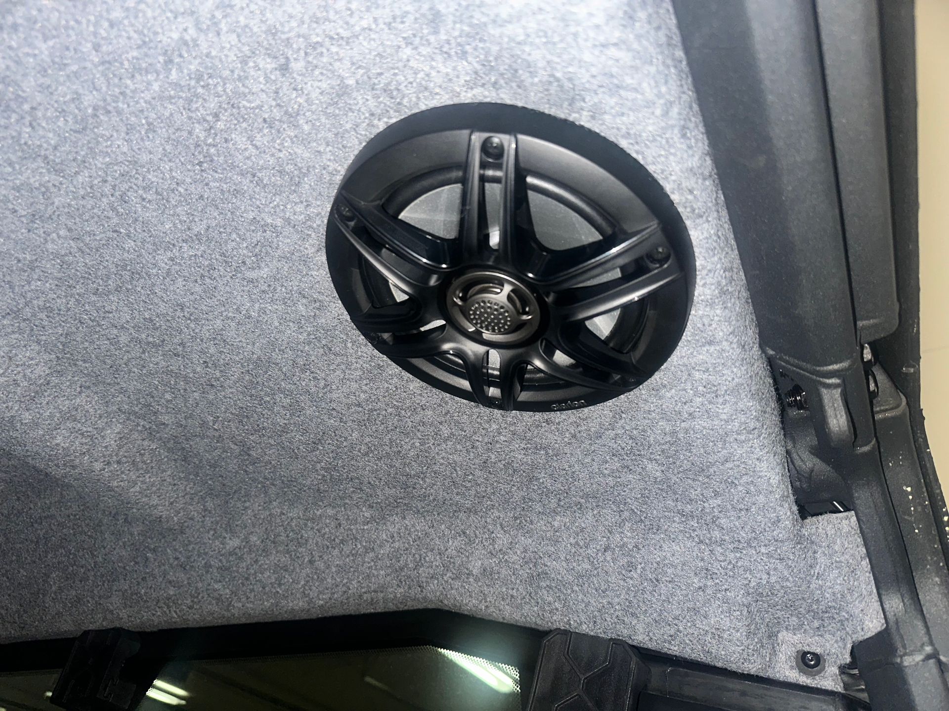 Black speaker mounted on a gray, carpeted car ceiling.