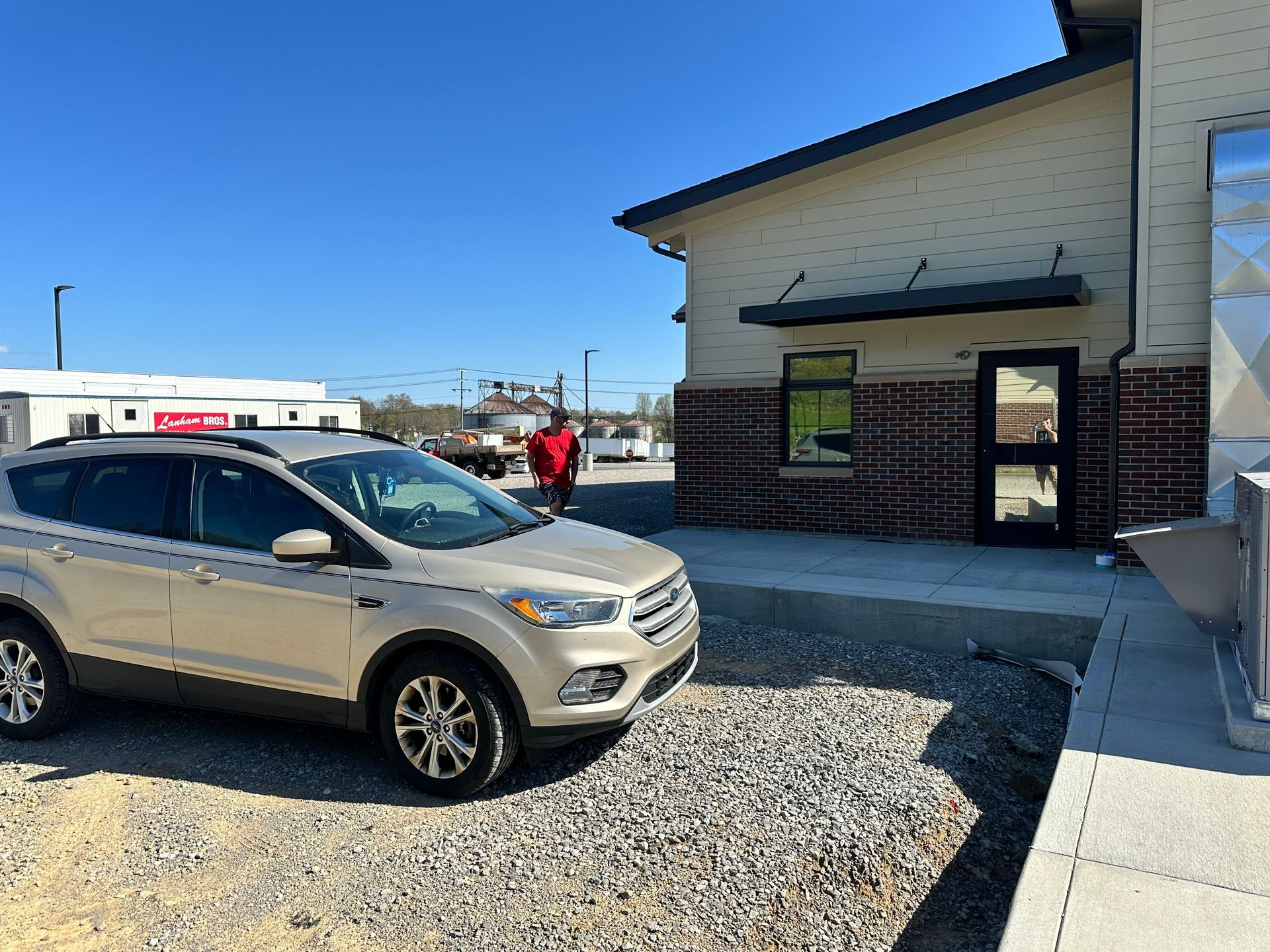 Gold SUV parked by a building; person in red shirt walks in the background. Sunny day.