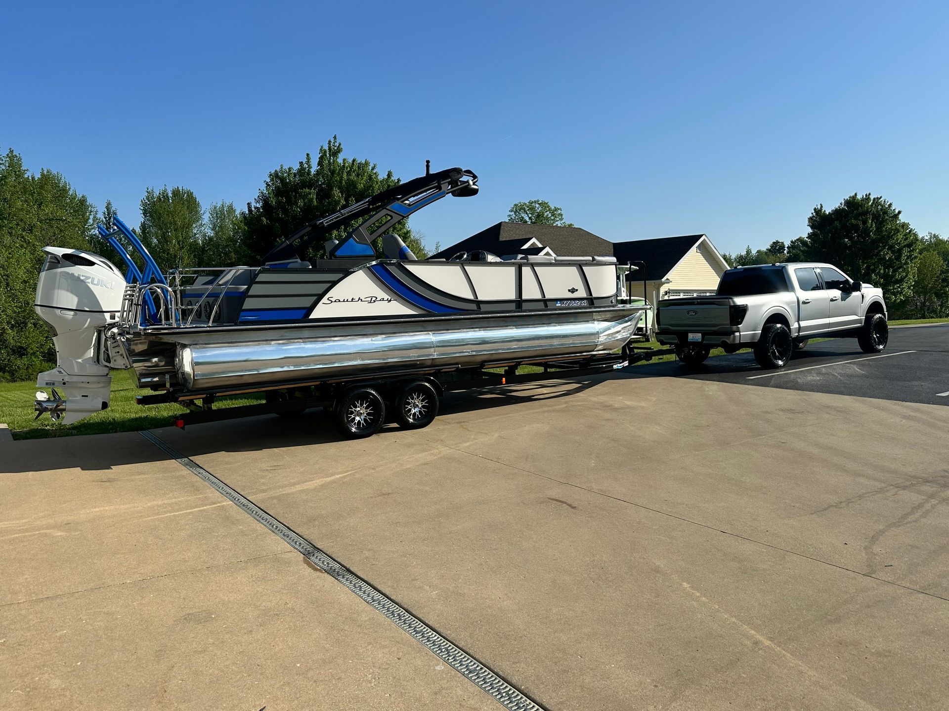 A pontoon boat on a trailer, towed by a pickup truck, parked in front of a house on a sunny day.