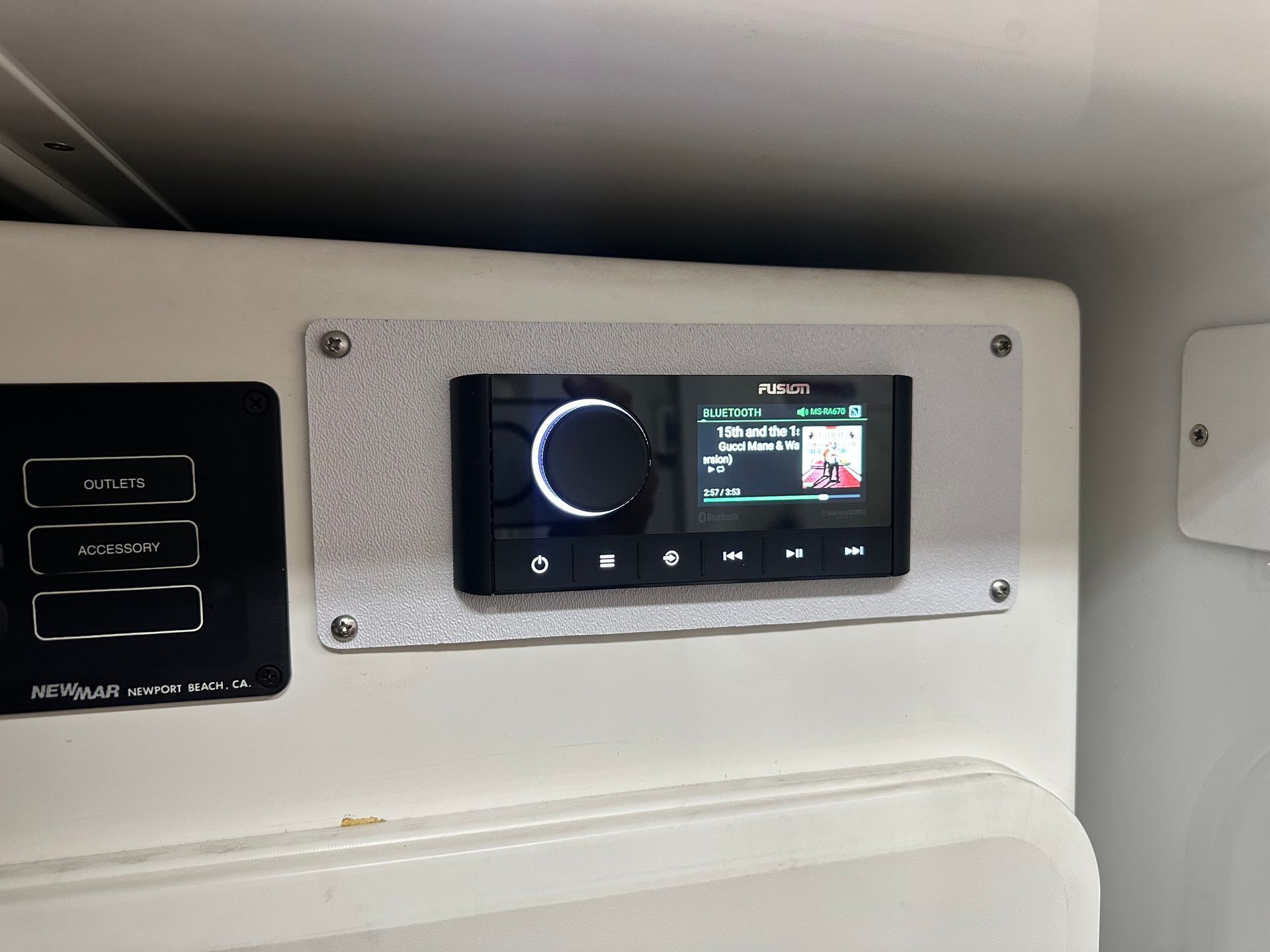 A boat's stereo system mounted on a white surface, with buttons and a screen displaying music information.