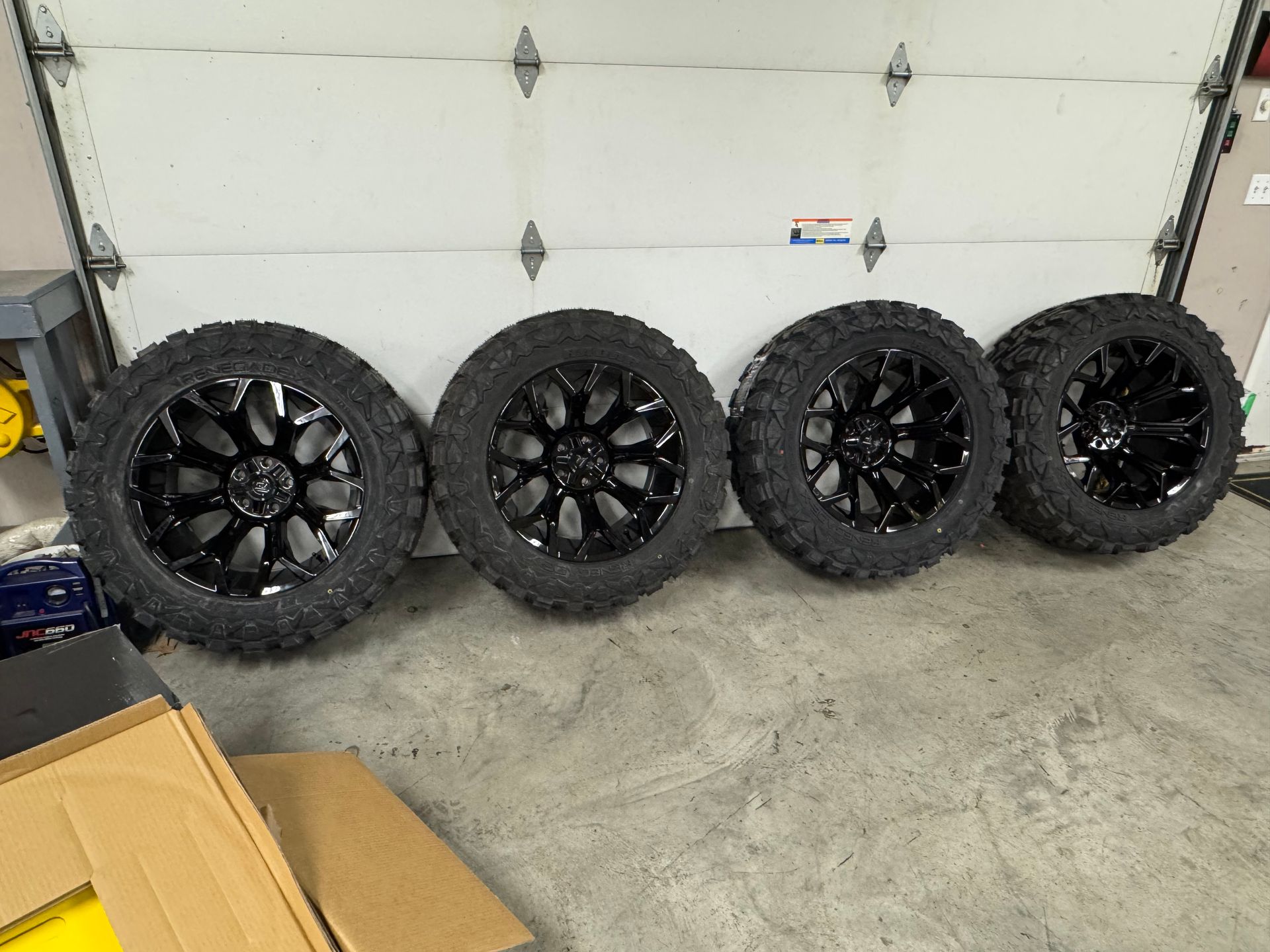 Four black off-road tires with black rims are set in front of a white garage door.