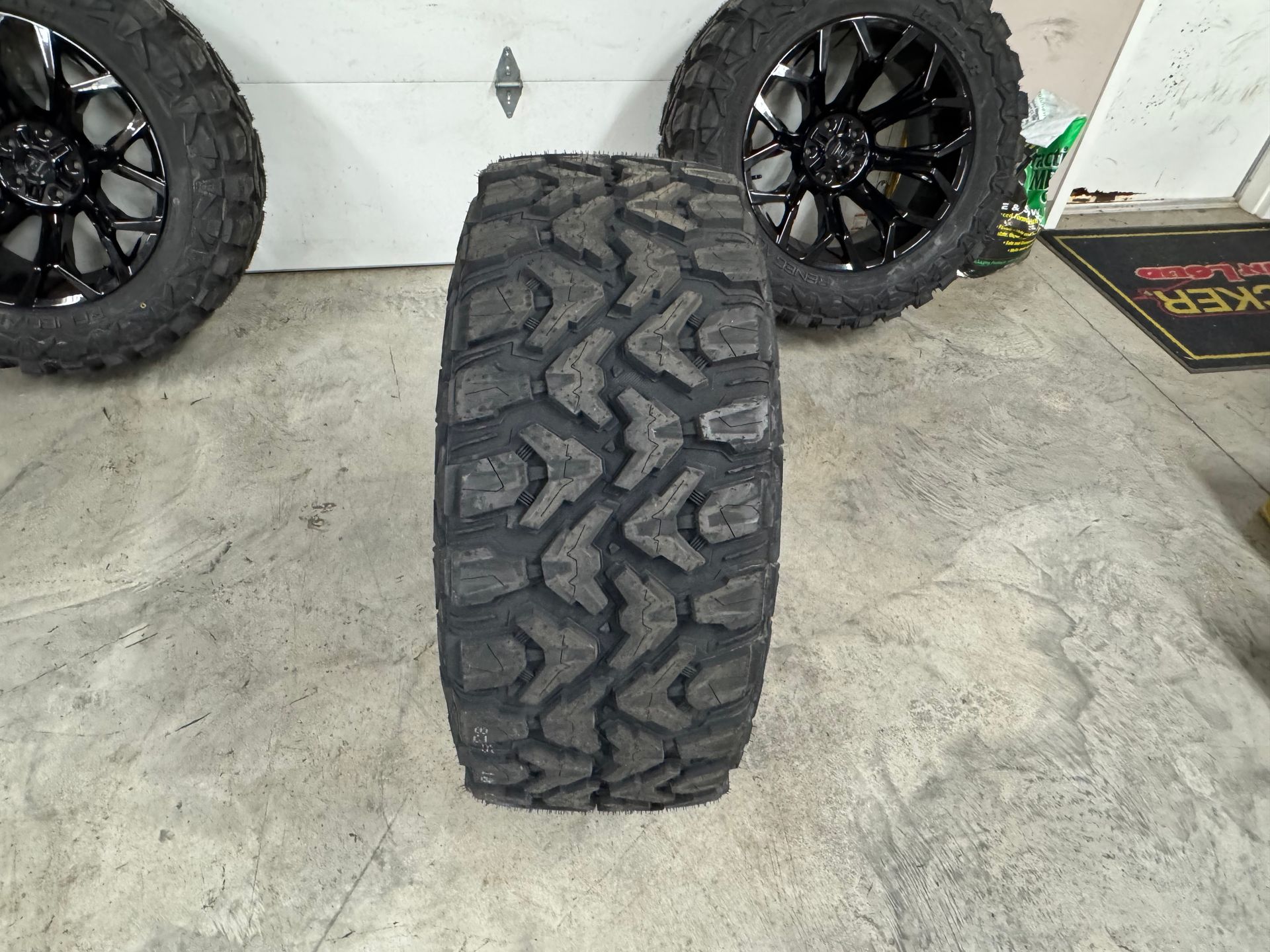 Off-road tire tread, with two black rims in background, on a concrete floor.