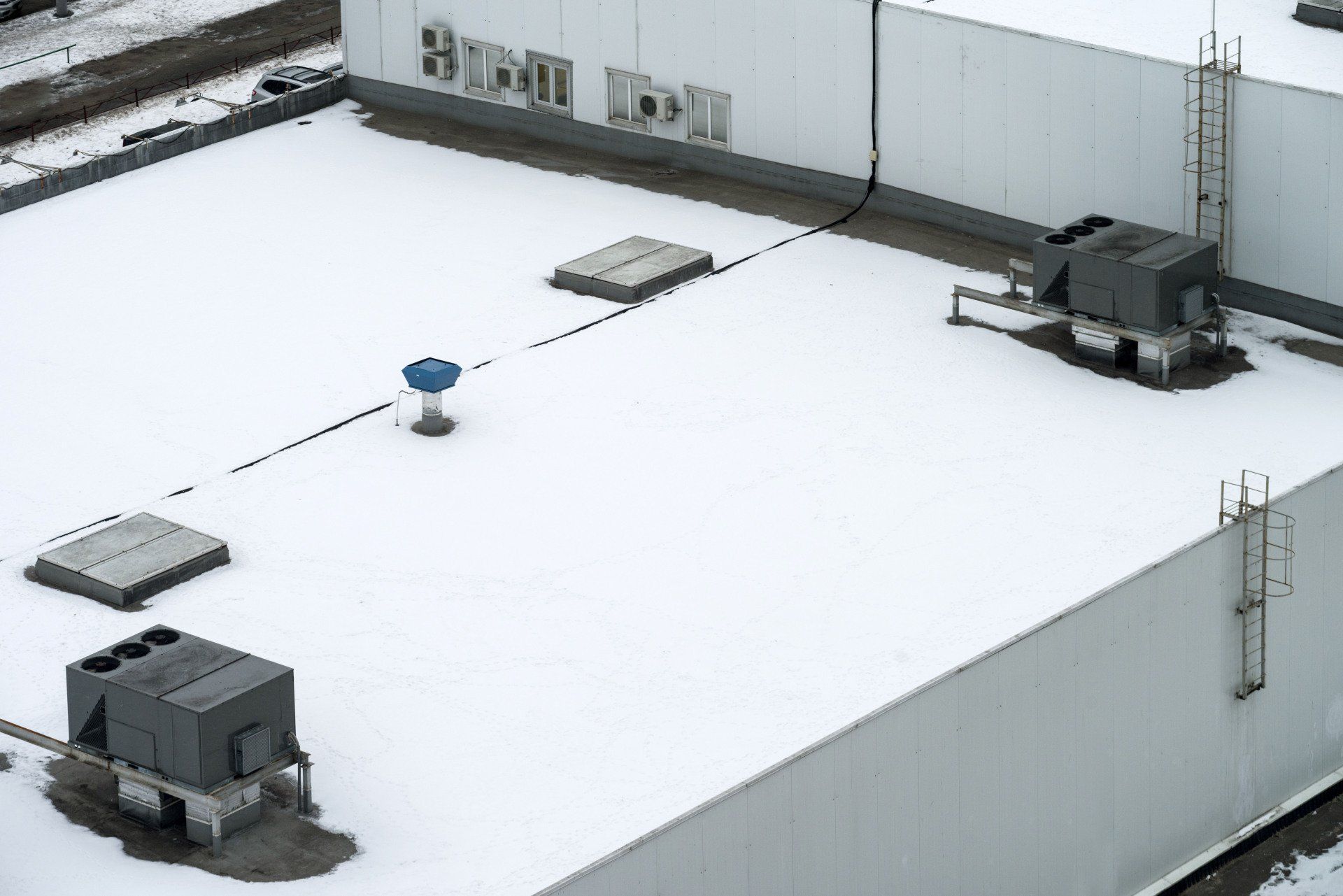 Snow-covered commercial building rooftop with HVAC units, hatches, and a ladder.