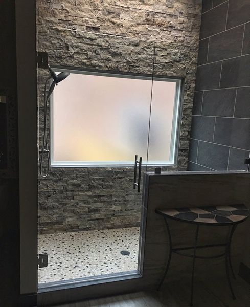 Shower with glass door and frosted window, stone and gray tile walls. A decorative table is near the shower.
