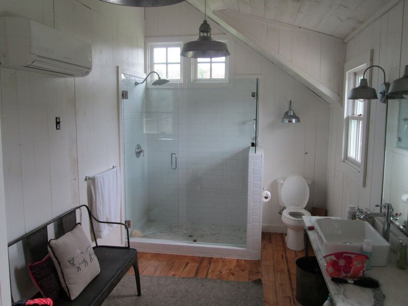White bathroom with a large glass shower, toilet, and vanity. A bench sits near the shower, with wood flooring and an angled ceiling.