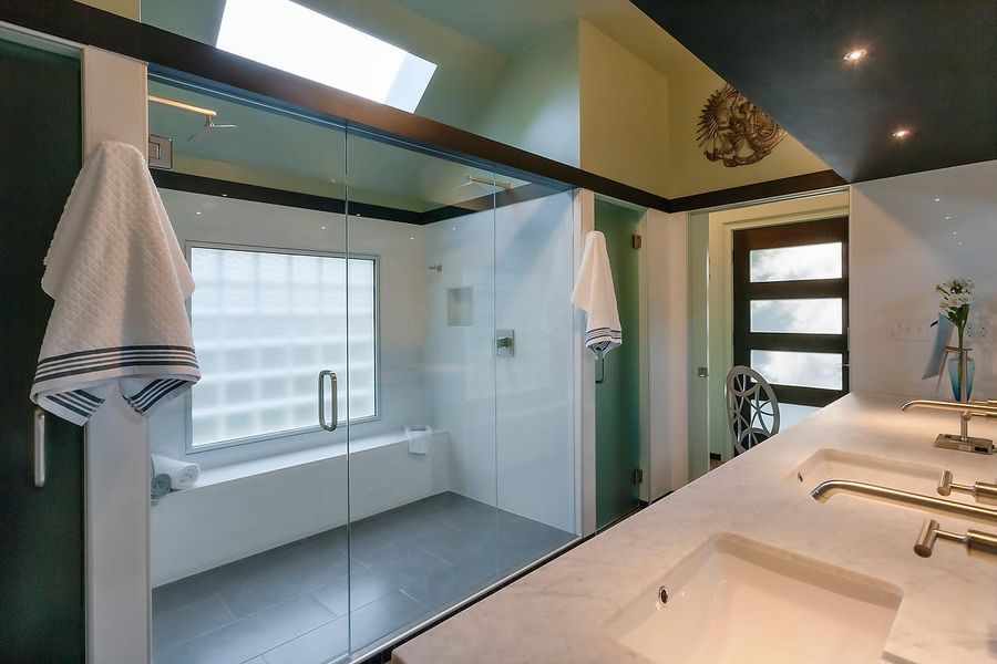 Modern bathroom with glass shower, dual sinks, and a window with light-blocking glass. Dark accents and green walls.