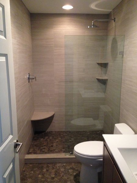 A small, modern bathroom with a pebble-floored shower, glass door, toilet, and sink. The walls are tiled, and the door is open.