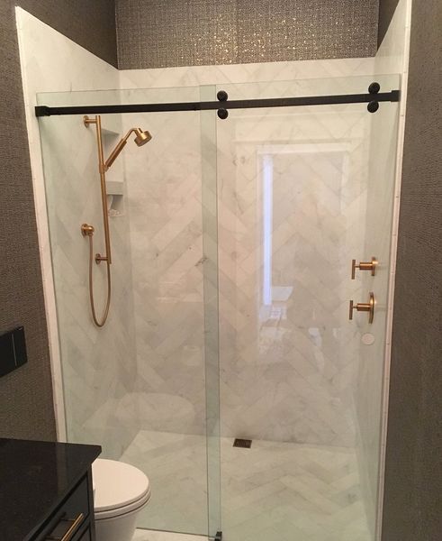 A modern shower with a glass sliding door, gold fixtures, and white marble tile with a herringbone pattern. The walls are dark textured gray.