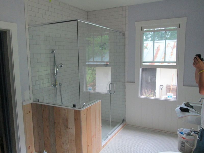 Bathroom with glass shower enclosure, wood-paneled base, white tile walls, and window with white trim and blue walls.