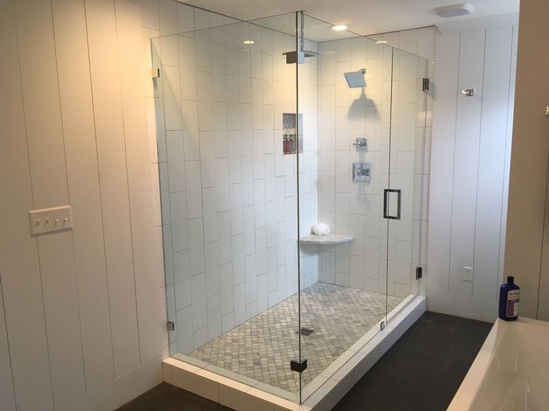 A modern glass shower enclosure with white tiled walls and a stone floor, featuring a built-in bench and a showerhead.