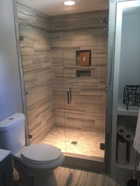 A modern bathroom with a glass-enclosed shower, toilet, and built-in shelving with towels and toilet paper. Light gray and beige tones.