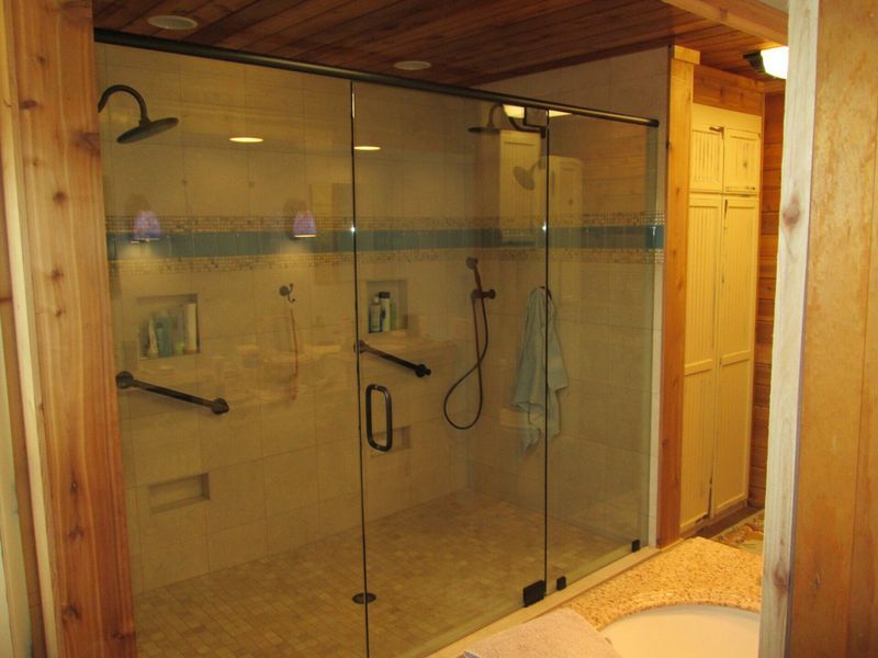 Glass-walled shower with grab bars and a handheld showerhead.  The bathroom has light-colored tiled walls and wood trim.