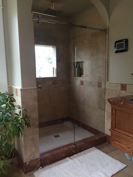 A bathroom with a tiled shower, a glass door, and a red stone threshold. The walls are tan, and a white rug is on the floor.