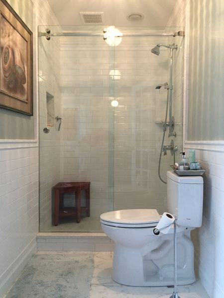 A small bathroom with a toilet, shower, and wooden stool. White tile and wainscoting contrast with a framed picture and vertical stripe wallpaper.