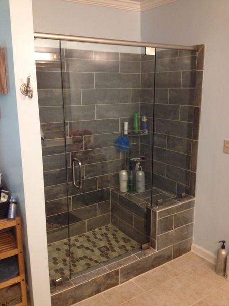A modern bathroom shower with gray tile walls, a glass door, and a built-in bench, with various toiletries visible.