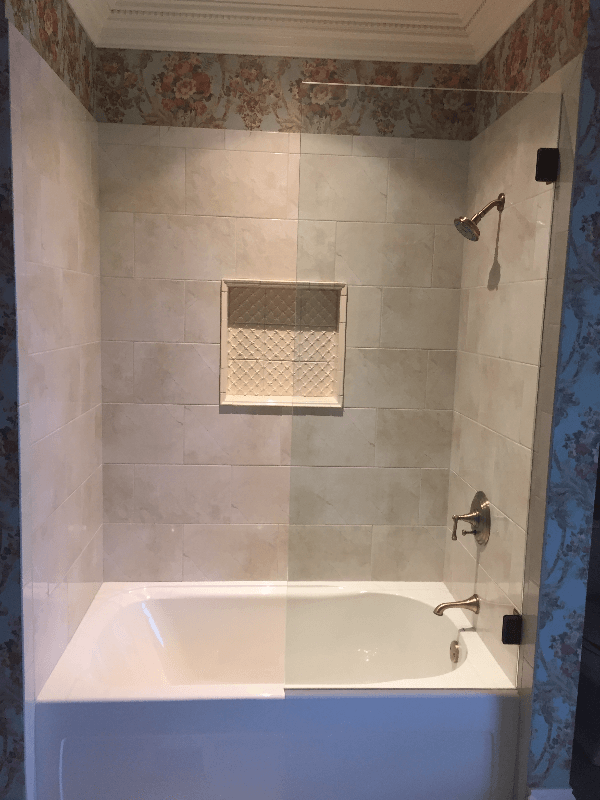 A tiled shower/tub combo with a glass door, showerhead, and built-in shelf; the background features wallpaper with a floral design.