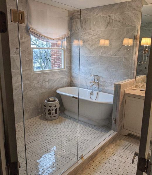 Bathroom with a glass shower, a white clawfoot tub, and gray tiled walls. A white stool sits near the window.