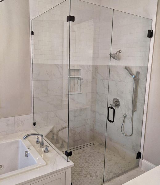 A glass-enclosed shower with black hardware and white marble walls and floor. There's a bathtub in front of the shower.