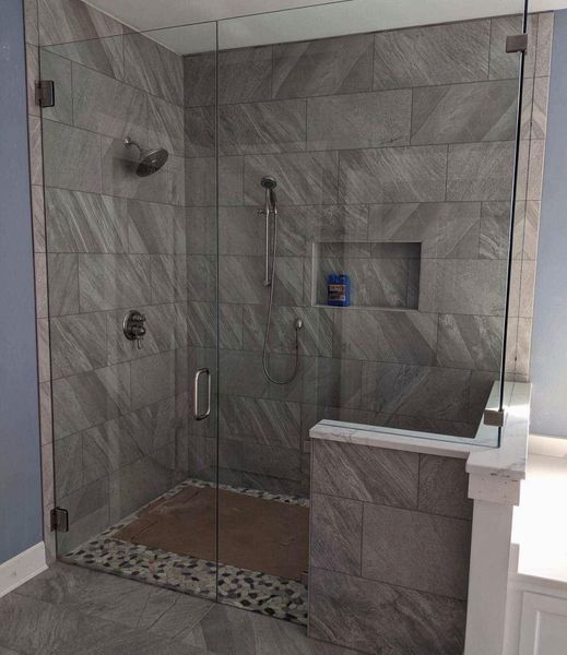 A modern shower enclosure with gray tile walls, glass doors, and a pebble floor. It features a showerhead, handheld shower, and built-in shelf.
