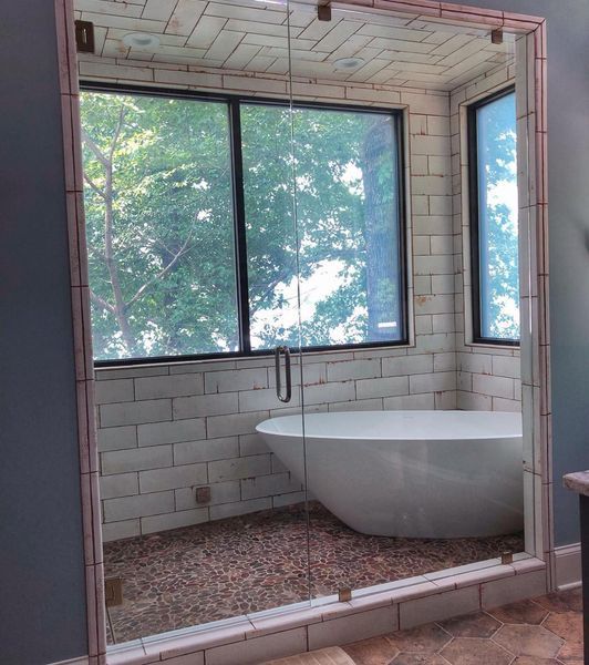 A modern bathroom with a white bathtub and a glass shower enclosure.  A window offers a view of green trees, with tile and pebble flooring.