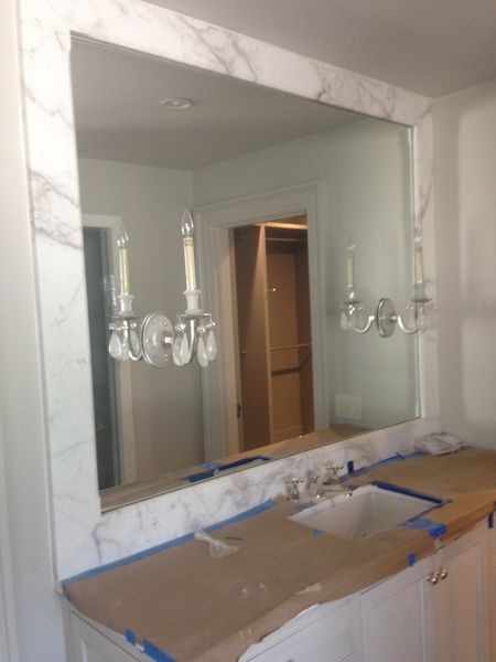 Bathroom with a large mirror framed in white marble, reflecting a closet and wall sconces. The vanity is white with a marble countertop.