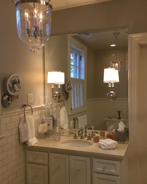 Bathroom with large mirror, vanity, and light fixtures. White cabinets and subway tile, with a window and shutters visible in the mirror.