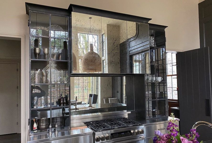 Kitchen with a mirrored backsplash, stove, shelves, and a dark gray cabinet. The kitchen has stainless steel elements and a window in the background.