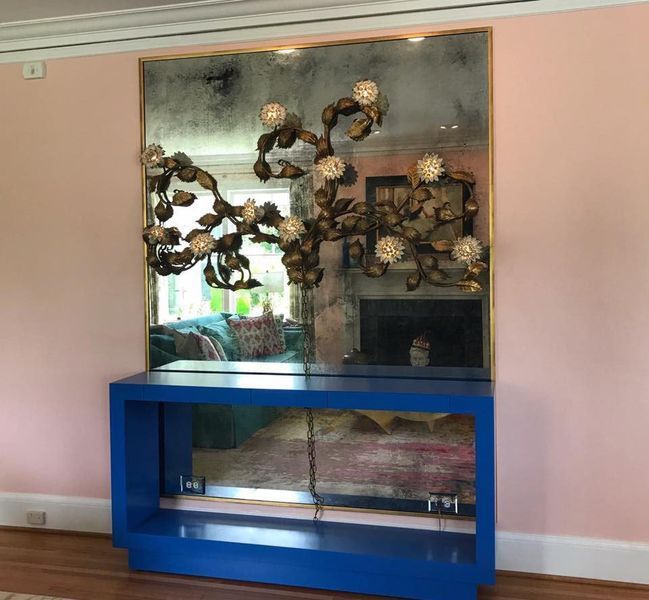 A large mirror with a gold floral design sits above a bright blue console table in a pink room.