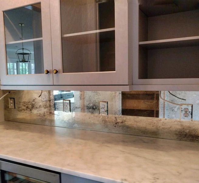 Kitchen with distressed mirror backsplash, light gray cabinets, and a marble countertop. Gold cabinet knobs and electrical outlets are visible.