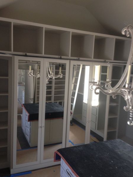 A partially built walk-in closet with mirrored sliding doors, shelving, and a central island. Soft light streams in from a window.
