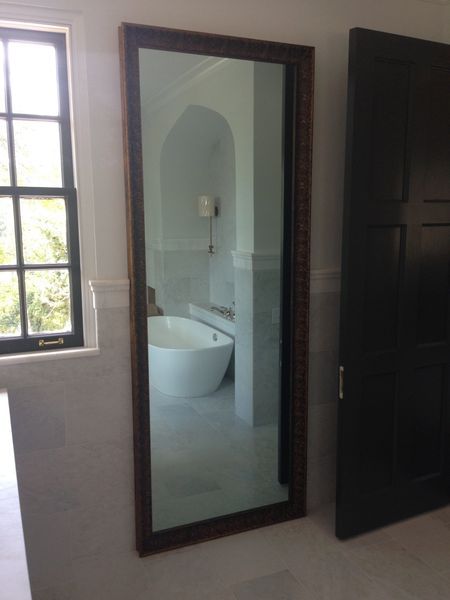 A long, rectangular mirror with a brown ornate frame reflecting a white bathroom with a bathtub. A black door and window are to the right and left.