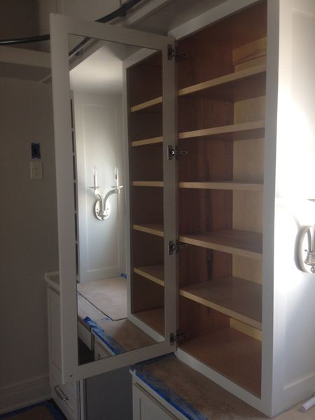 White bathroom cabinet with mirror door open, revealing shelves. The mirror reflects the room, including sconces on the wall.