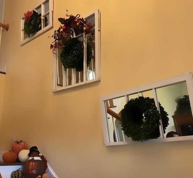 Yellow wall decorated with three faux window frames holding wreaths. Pumpkins and a turkey figurine sit on a staircase.