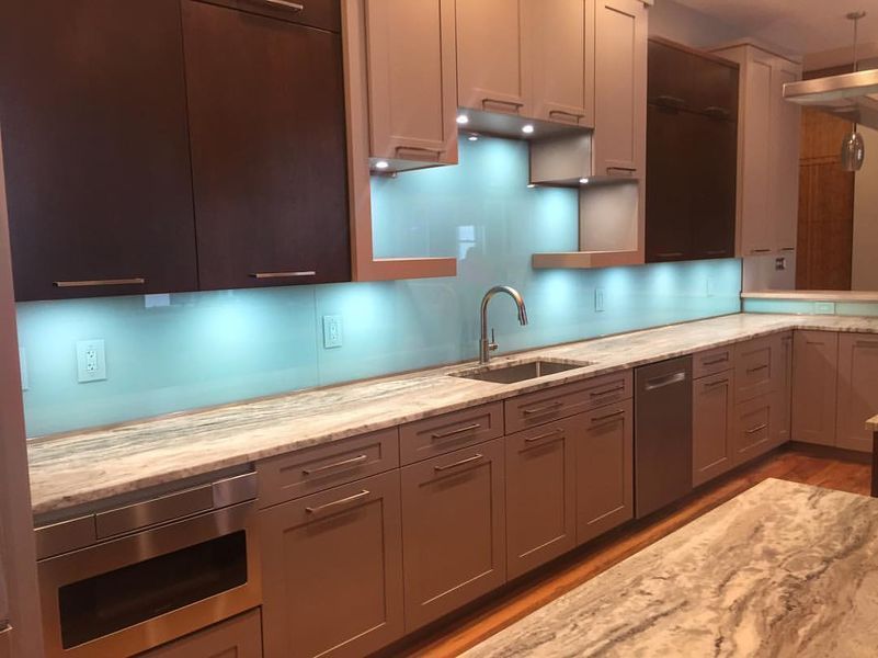 Modern kitchen with light gray cabinets, a light blue glass backsplash, and dark brown upper cabinets. Stainless steel appliances and a marble countertop are present.