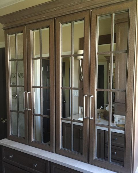 A built-in cabinet with four glass-paned doors, reflecting a bathroom interior. The cabinet is brown, with silver hardware.