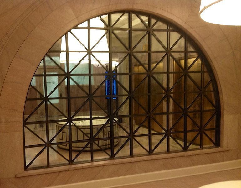 Arched window with black metal gridwork and clear glass, set in a light stone wall. A room with a circular object is visible through the window.