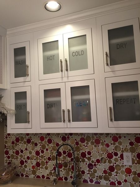 White kitchen cabinets with frosted glass doors labeled for laundry, above a red and beige mosaic backsplash and a sink.