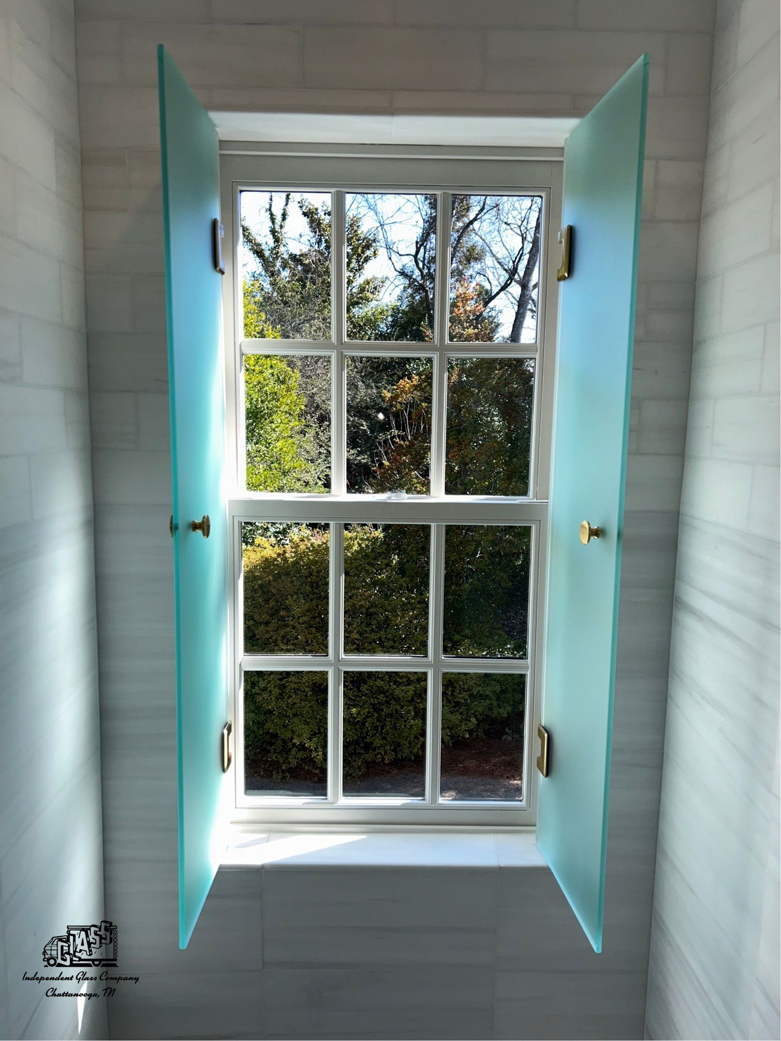 A white window with a grid design is flanked by open, frosted teal glass shutters. The window looks out onto a green, tree-filled outdoor view.