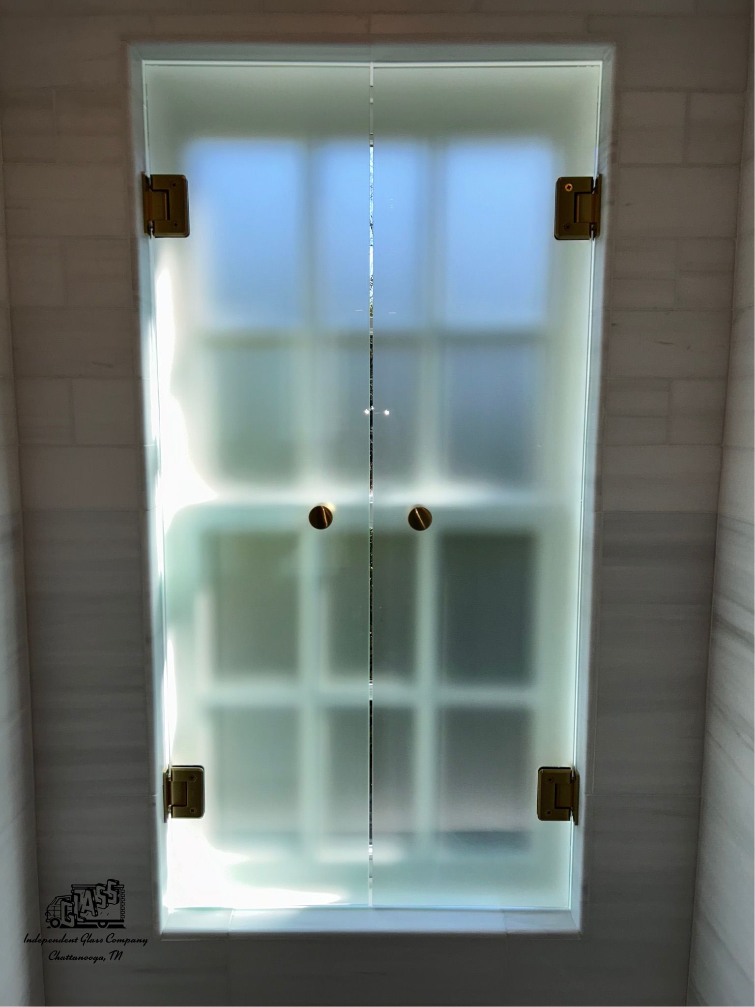 Frosted glass shower doors with brass hinges and handles, framed by light marble walls.