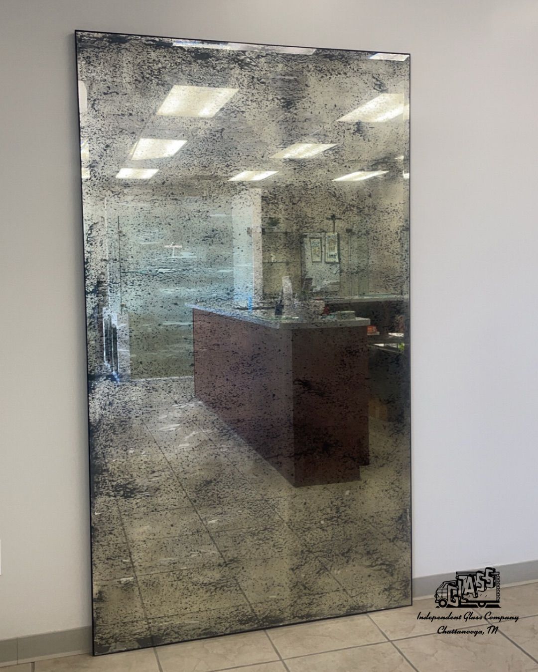 Large rectangular antique mirror leans against a white wall, reflecting a room with a wooden counter and overhead lights.