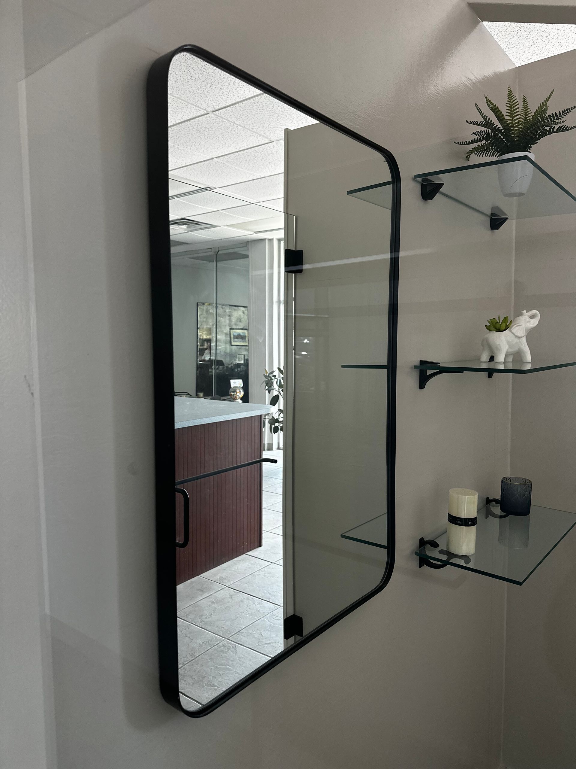 Black-framed mirror on a white wall, reflecting a room with brown cabinets and glass shelves with small plants.