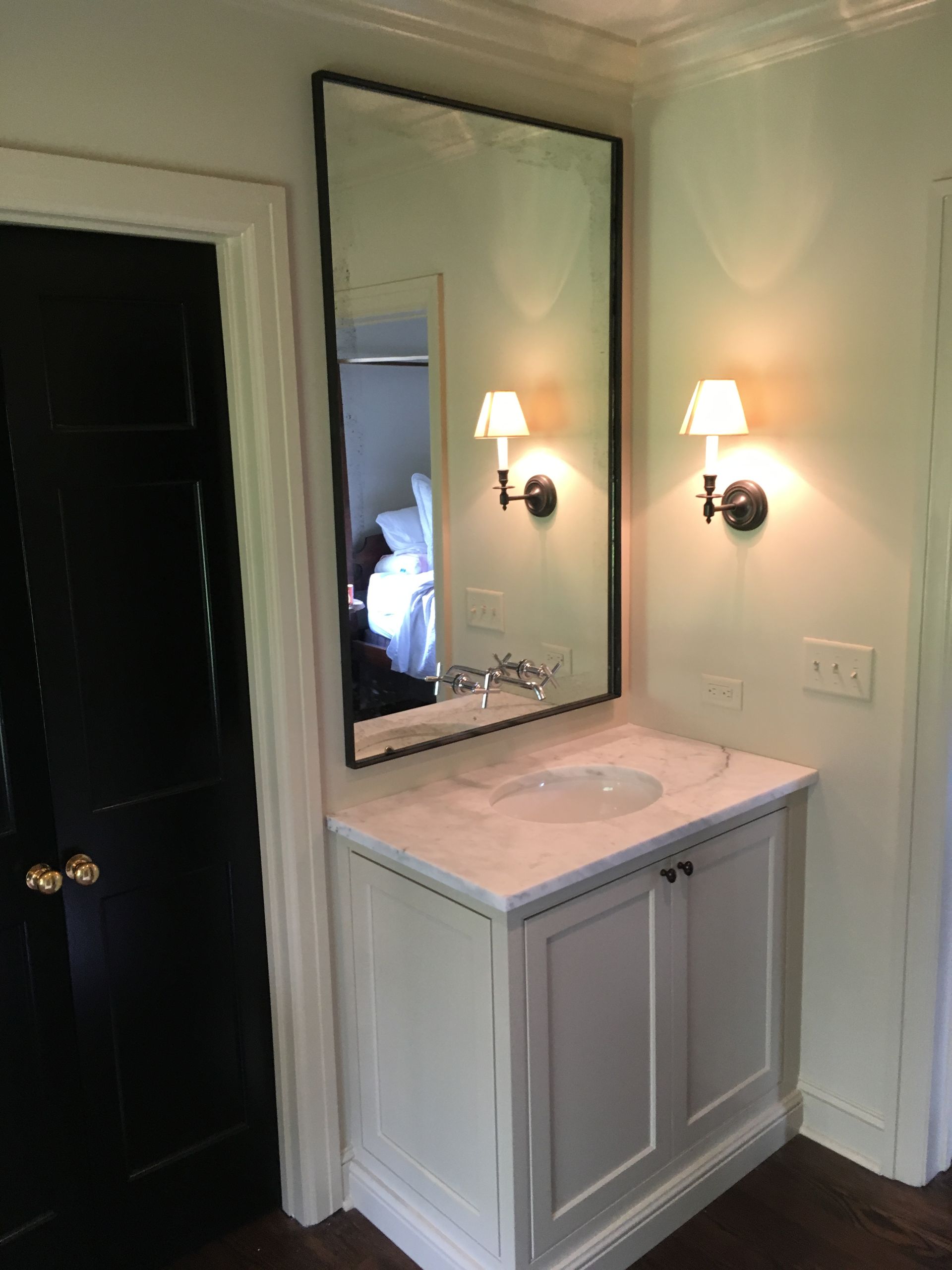 A bathroom with a white cabinet, marble countertop, black-framed mirror, and sconces. A black door is to the left.