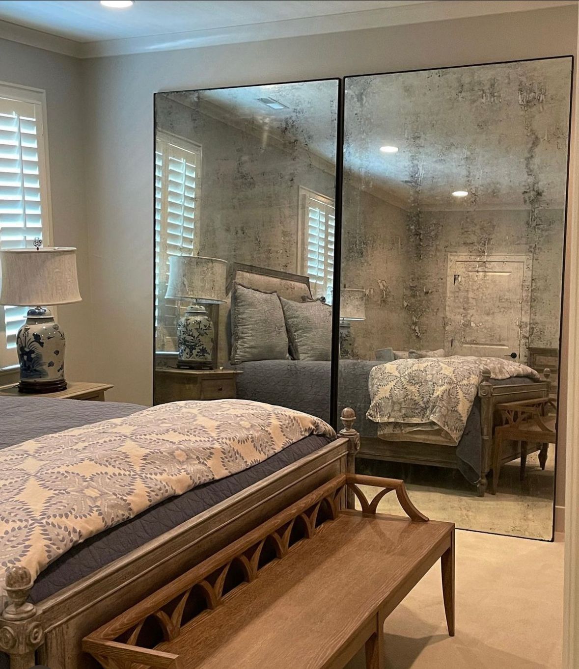 Bedroom with large, antique-style mirrored closet doors reflecting a bed, windows with shutters, and bedside tables.