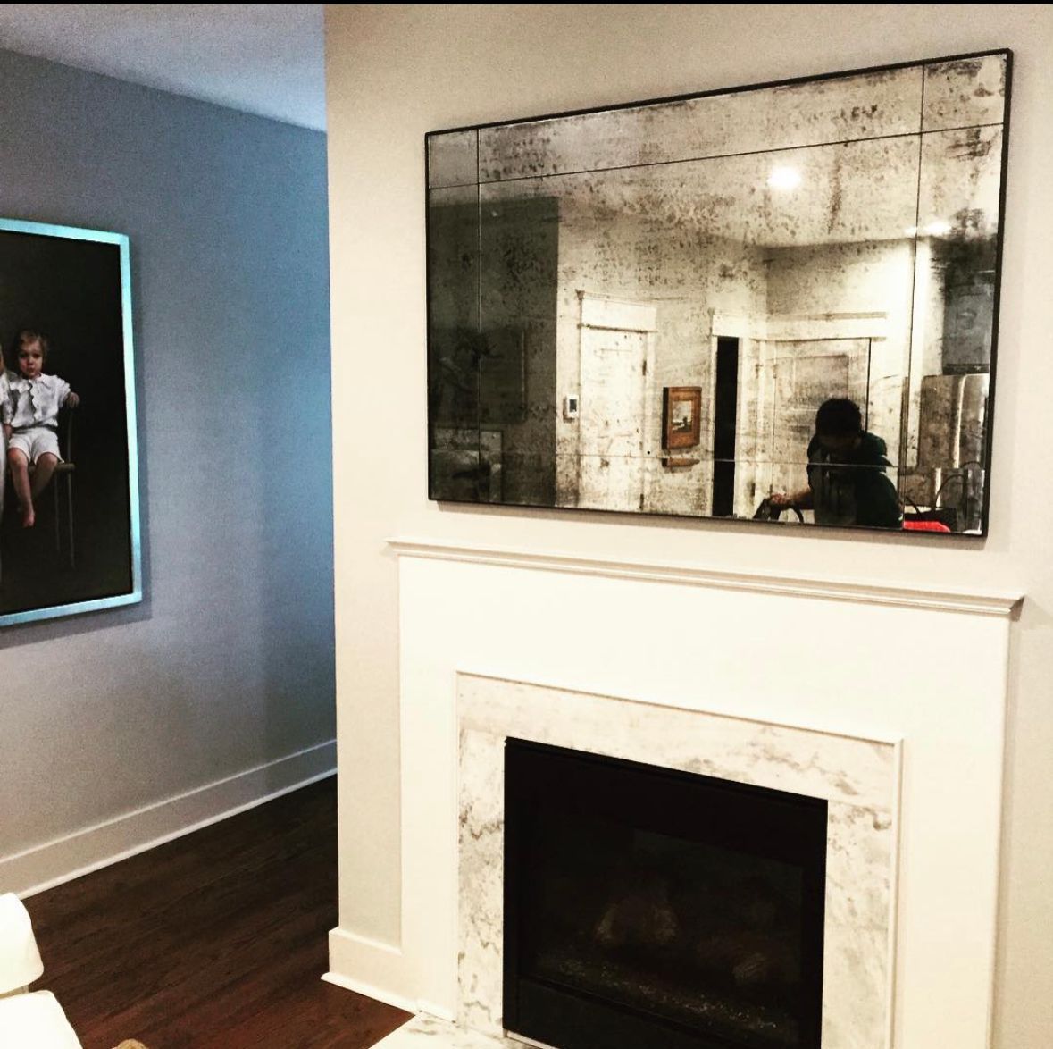 Living room with fireplace and large antique mirror. Artwork on the left. Dark wood floors, light walls.