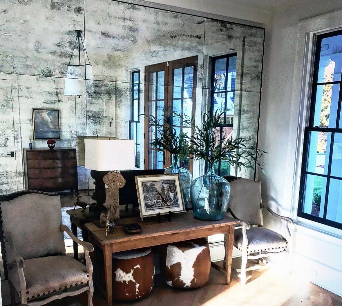 Living room with a distressed mirror wall, wooden furniture, and large windows. Features include two chairs, a table with a lamp and decor, and plants.