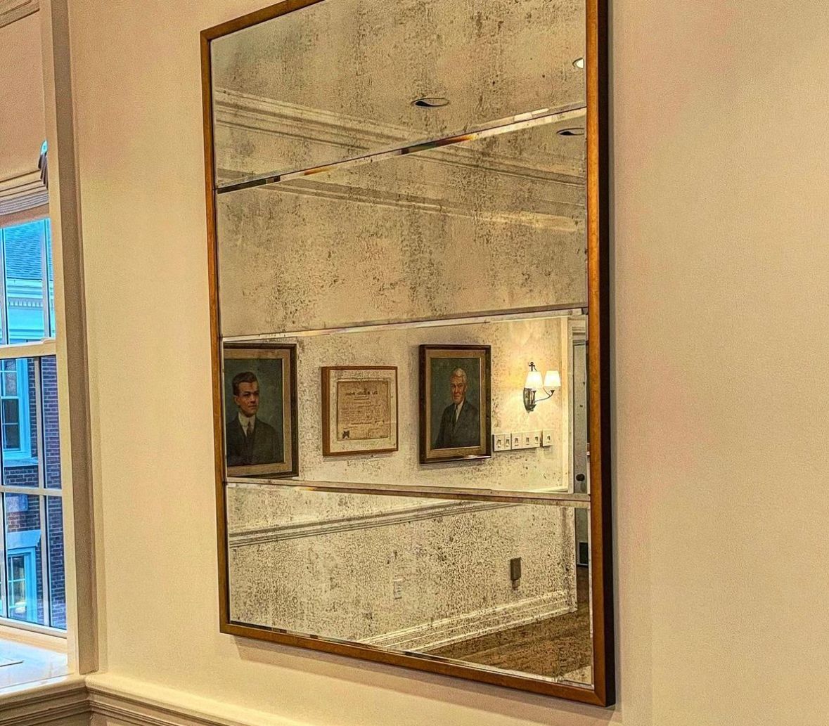 Large antique mirror reflecting a room with portraits, staircase, and a window with a partially visible building. Mirror has a gold frame.