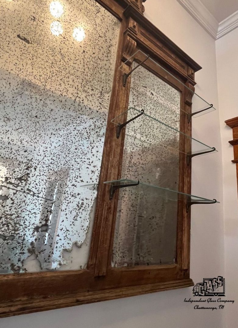 Large antique mirror with wooden frame, glass shelves, and a distressed, speckled appearance. Mounted on a white wall.