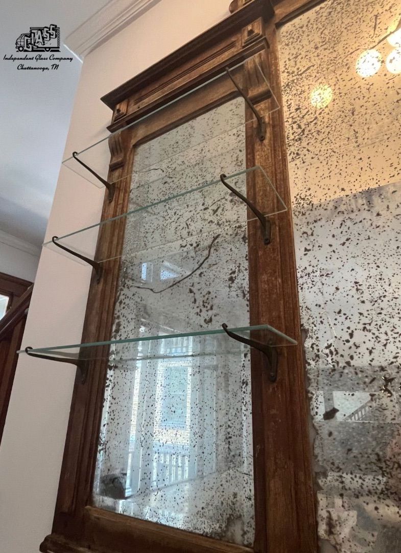 Large, antique mirror with a distressed finish, wooden frame, and glass shelves attached. The mirror reflects a bright room.