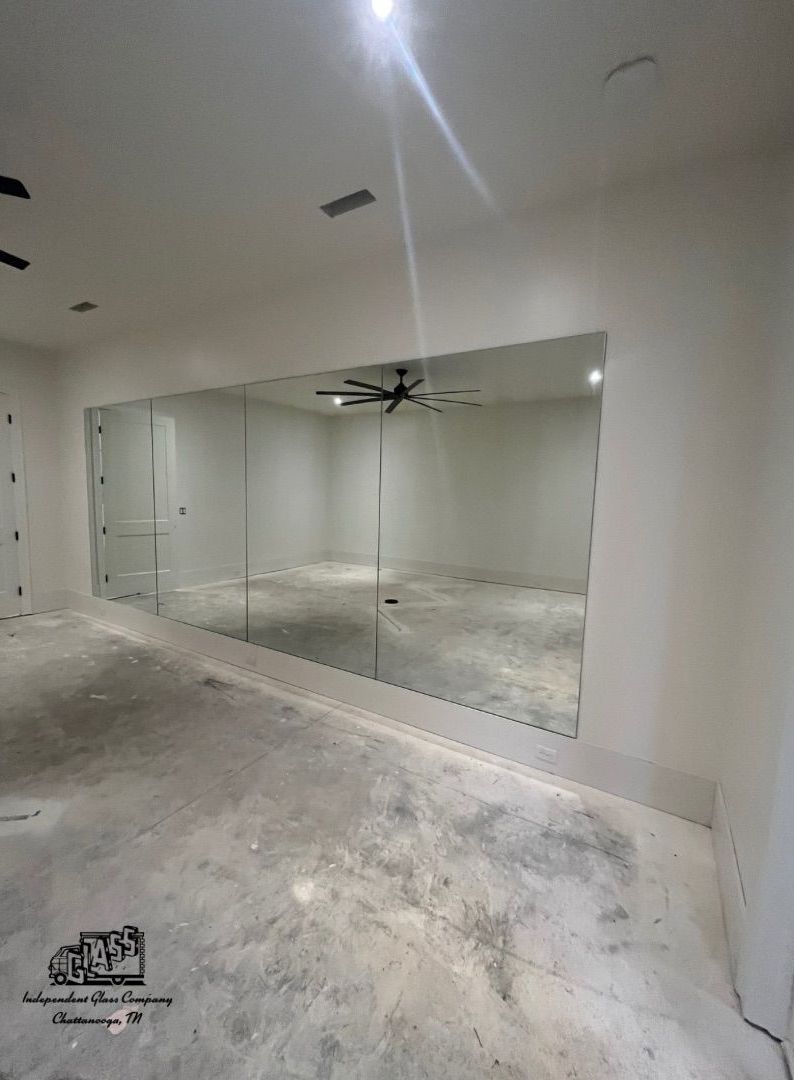 A large, rectangular mirror covers most of a white wall in an unfinished room. The mirror reflects the room, including a ceiling fan.