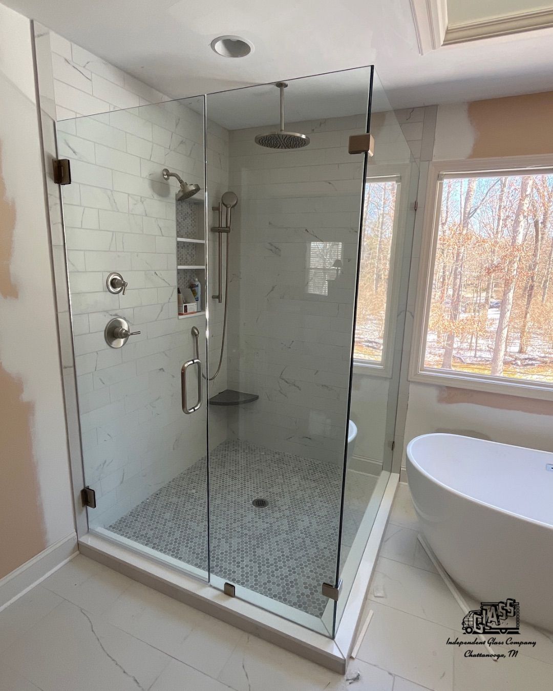 A modern bathroom with a glass-enclosed shower, white marble walls, and a freestanding tub next to a window overlooking a snowy scene.